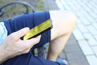 Bir elinde akıllı bir telefon olan biri, yeşil alanlarla çevrili bir bankta oturuyor. Telefondan yansıyan güneş ışığı rahatlatıcı bir atmosfer yaratıyor..