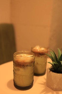 Buzlarla dolu iki ferahlatıcı içecek basit bir masada dinleniyor. Yumuşak sıcak ışık, yakınlardaki küçük bir bitki tarafından tamamlanmış rahat bir atmosfer yaratır. Rahatlamak ya da sosyalleşmek için ideal.