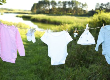 Farklı renklerde bebek kıyafetleri, güzel bir açık hava ortamında çamaşır ipinde kuruyor. Huzurlu bir nehir ve yeşillik sahneyi çevreleyerek sakin bir atmosfer yaratıyor..