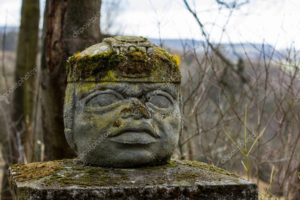 The Aztec statue on a pylon. The head or face is made from stone and covered with moss. 