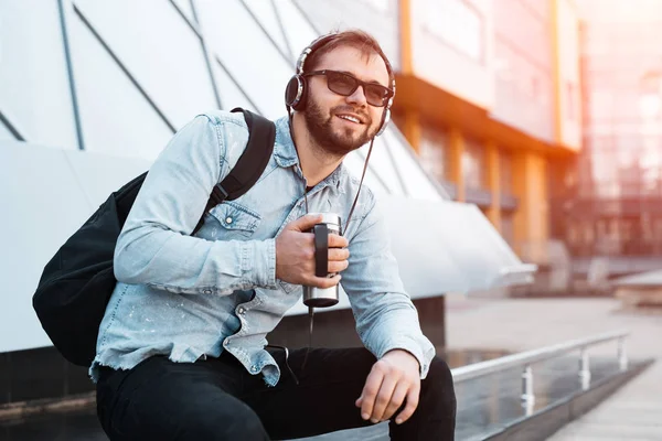 Moderno hipster barbudo sonriente con mochila negra, auriculares en las orejas y taza termo con café en la mano — Foto de Stock