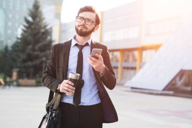Hipster etkilendim işadamı kahve termo kupa el ve siyah çanta ofis yapı zemin üzerine Smartphone kullanarak.