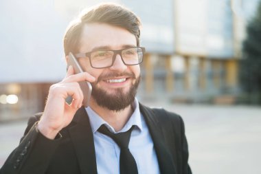 Ofis yapı ve güneş ışığı arka plan üzerinde smartphone kullanarak takım elbiseli gülümseyen sakallı işadamı portresi.