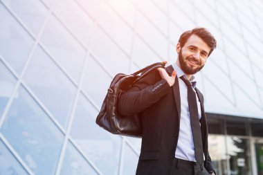 Gülümseyen hipster portresi işadamı gökdelen windows arka plan üzerinde çanta ile takım elbiseli sakallı.