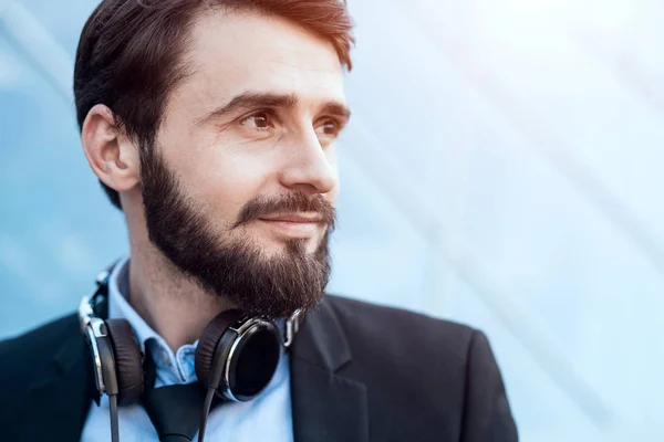 Close-up portrait of bearded hipster with headphones on neck in suit, on windows skyscraper background.