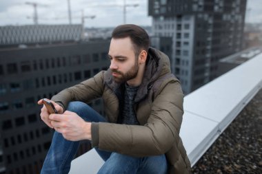 Yüksek bir binanın çatısında akıllı telefon, sosyal ağ, sakallı hippi adam bakar.