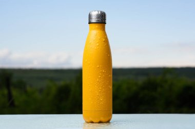 Termo Paslanmaz şişe, su ile püskürtülür. Gökyüzü ve orman arka plan üzerinde. Cam masanın üzerinde. Termos mat sarı renk