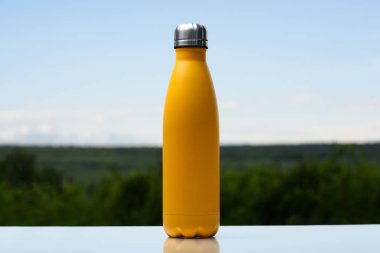 Termo Paslanmaz şişe su, çay veya kahve için. Gökyüzü ve orman arka plan üzerinde. Cam masanın üzerinde. Termos mat sarı renk