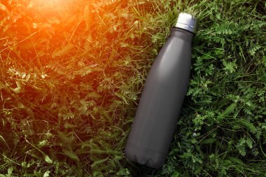 Paslanmaz termos su şişesi, mat siyah renk. Güneş ışığı etkisi yeşil çim zemin üzerine izole mockup