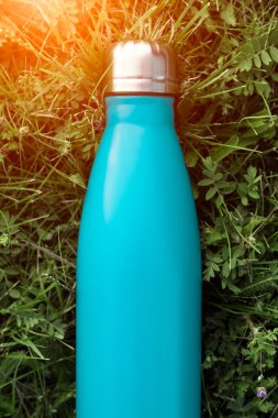 Paslanmaz termos su şişesi, açık mavi renk. Güneş ışığı etkisi yeşil çim zemin üzerine izole