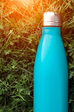 Paslanmaz termos su şişesi, açık mavi renk. Güneş ışığı etkisi yeşil çim zemin üzerine izole