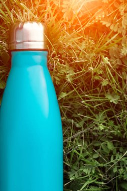 Paslanmaz termos su şişesi, açık mavi renk. Güneş ışığı etkisi yeşil çim zemin üzerine izole