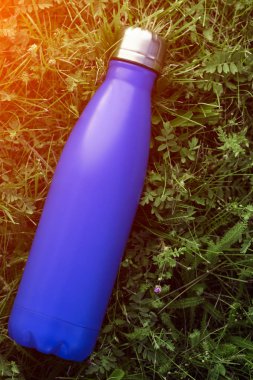 Paslanmaz termos su şişesi, mat mavi renk. Güneş ışığı etkisi yeşil çim zemin üzerine izole mockup