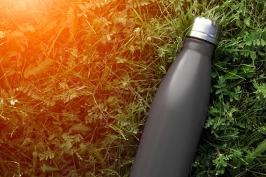 Paslanmaz termos su şişesi, mat siyah renk. Güneş ışığı etkisi yeşil çim zemin üzerine izole mockup
