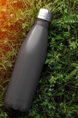 Paslanmaz termos su şişesi, mat siyah renk. Güneş ışığı etkisi yeşil çim zemin üzerine izole mockup