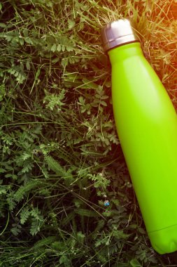 Paslanmaz termos su şişesi, mat açık yeşil renk. Güneş ışığı etkisi yeşil çim zemin üzerine izole mockup