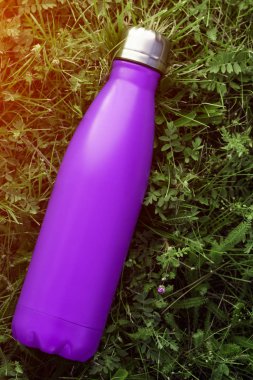 Paslanmaz termos su şişesi, mat mor renk. Güneş ışığı etkisi yeşil çim zemin üzerine izole mockup