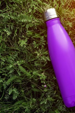 Paslanmaz termos su şişesi, mat mor renk. Güneş ışığı etkisi yeşil çim zemin üzerine izole mockup