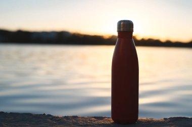 Paslanmaz kırmızı termo şişe su için. Günbatımı ve arka plan üzerinde su
