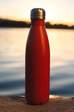 Paslanmaz kırmızı termo şişe su için. Günbatımı ve arka plan üzerinde su