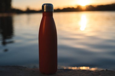 Paslanmaz kırmızı termo şişe su için. Günbatımı ve arka plan üzerinde su
