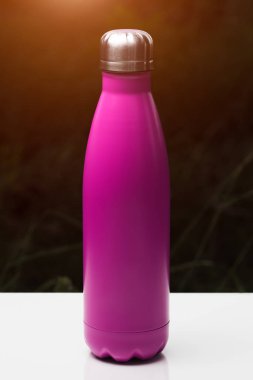 Paslanmaz termo şişe su, çay ve kahve, beyaz masada. Güneş ışığı etkisi ile karanlık çim arka plan. Termos pembe renk