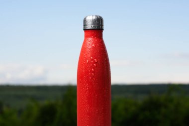 Termo Paslanmaz şişe, su ile püskürtülür. Gökyüzü ve orman arka plan üzerinde. Termos mat kırmızı renk