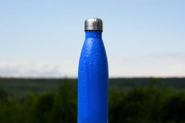 Termo Paslanmaz şişe, su ile püskürtülür. Gökyüzü ve orman arka plan üzerinde. Termos mat mavi renk