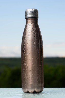 Termo Paslanmaz şişe, su ile püskürtülür. Gökyüzü ve orman arka plan üzerinde. Cam masanın üzerinde. Termos maro renk