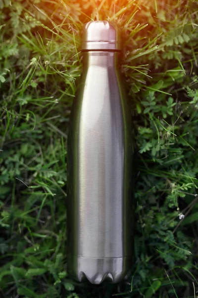 Stainless thermos water bottle, blue color. Mockup isolated on green grass background with sunlight effect. Glossy aluminum vacuum thermo tumbler flask