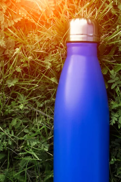 Stainless thermos water bottle, blue color. Mockup isolated on green grass background with sunlight effect. Glossy aluminum vacuum thermo tumbler flask