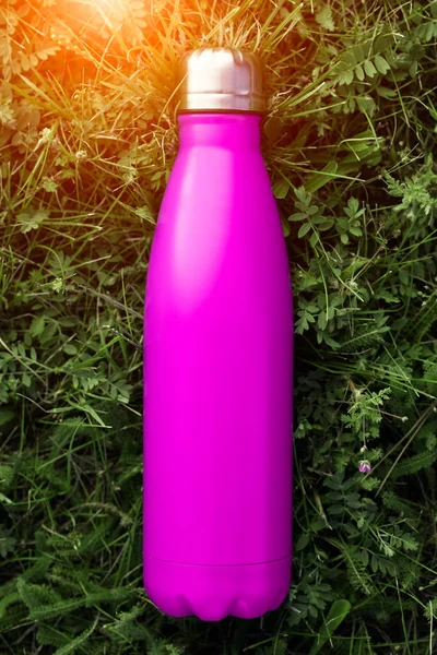 Stainless thermos water bottle, pink color. Mockup isolated on green grass background with sunlight effect. Glossy aluminum vacuum thermo tumbler flask