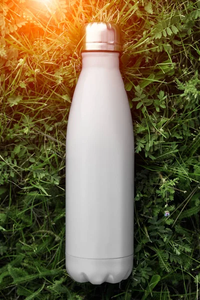 Stainless thermos water bottle, white color. Mockup isolated on green grass background with sunlight effect. Glossy aluminum vacuum thermo white tumbler flask