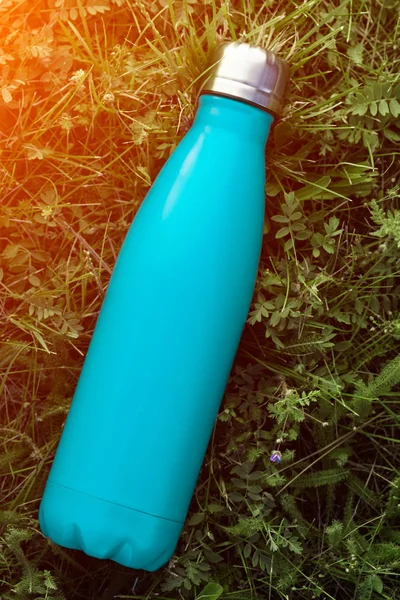 Paslanmaz termos su şişesi, mat açık mavi renk. Güneş ışığı etkisi yeşil çim zemin üzerine izole mockup