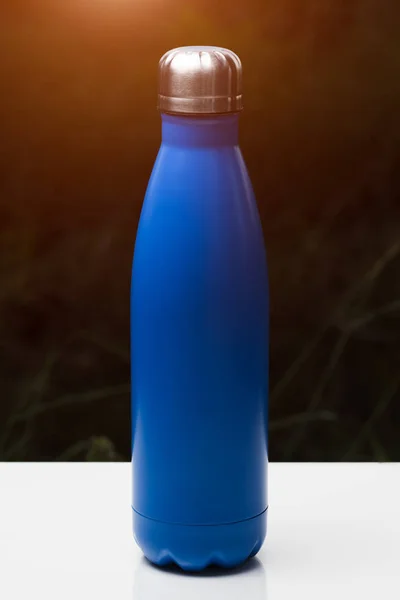 水やお茶 コーヒー 白いテーブルの上のステンレス熱ボトル 日光の影響で暗い草背景 魔法瓶ブルー色 — ストック写真