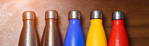 Colorful stainless thermo bottles, on a wooden table sprayed with water. Matte red bottle, blue, yellow and platinum color. With sunlight effect