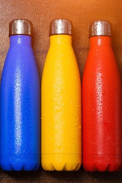 Colorful stainless thermo bottles, on a wooden table sprayed with water. Matte red bottle, blue, yellow and platinum color. With sunlight effect