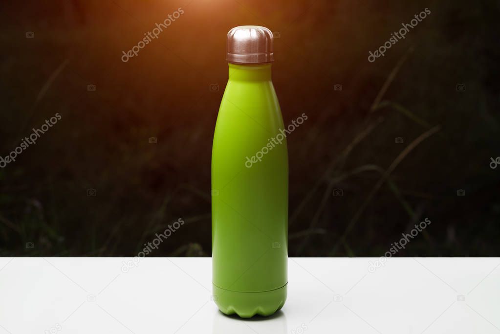 Stainless thermo bottle for water, tea and coffe, on white table. Dark grass background with sunlight effect. Thermos green color