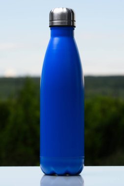 Termo Paslanmaz şişe su, çay veya kahve için. Gökyüzü ve orman arka plan üzerinde. Cam masanın üzerinde. Termos mat mavi renk