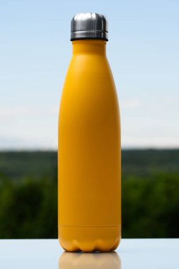 Termo Paslanmaz şişe su, çay veya kahve için. Gökyüzü ve orman arka plan üzerinde. Cam masanın üzerinde. Termos mat sarı renk