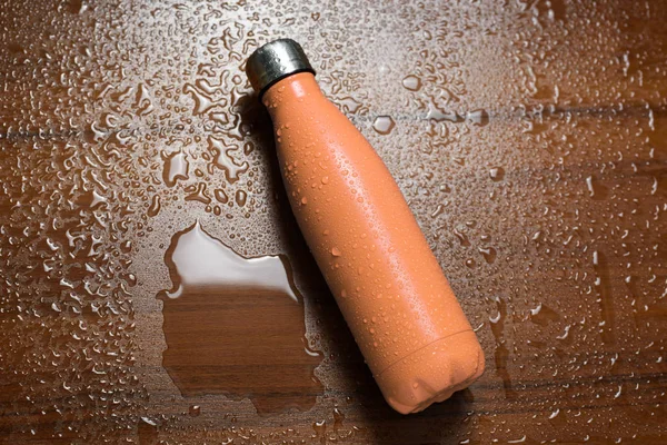 Stainless steel thermos bottle isolated on a wooden table sprayed with water. Orange color