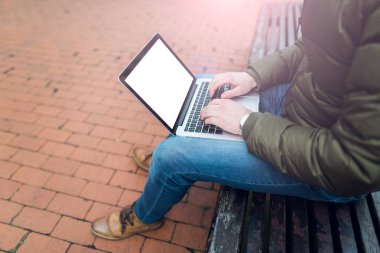 Park ahşap bir sandalyede oturan bir adamın elleri içinde laptop. Mavi jeans ve kahverengi ayakkabı giymiş. Boş beyaz ekran ile dizüstü mockup görüntü. Fotoğraf lens flare filtre etkisi ile..