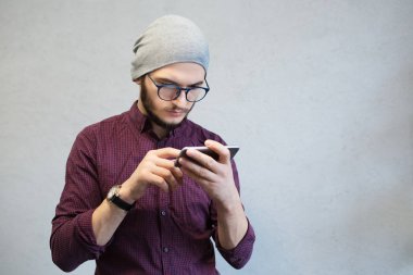 Dalgın teenaeger adam akıllı telefon, beyaz arka plan üzerinde kullanarak. Gömlek ve gümüş şapka, gözlük takan giymiş