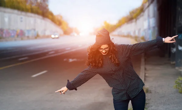 Glückliches, lockiges Teenager-Mädchen, das bei Sonnenuntergang auf der Straße steht. bekleidet mit Hemd und Hut, Brille. — Stockfoto