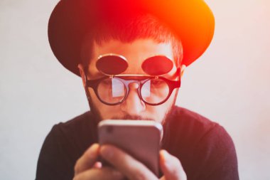 Sürpriz modern genç adam beyaz arka plan üzerinde smartphone kullanarak yakın çekim portre. Yenilikçi güneş gözlüğü ve siyah şapka giyiyor
