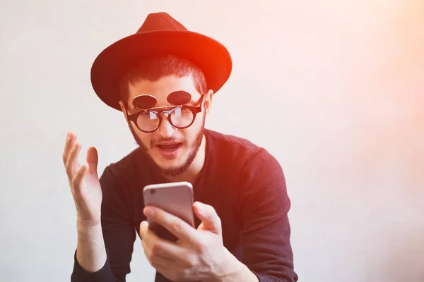 Portrait of emotional guy who using smartphone over white background, dressed in black. Wearing sunglasses and hat