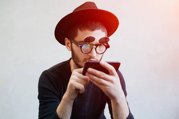 Portrait of young gamer guy with smartphone in hands over white background. Wearing sunglasses and hat