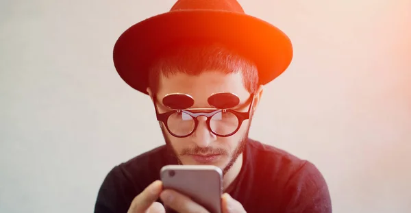 Close-up portrait of surprised modern young guy using smartphone over white background. Wearing hipster sunglasses and black hat