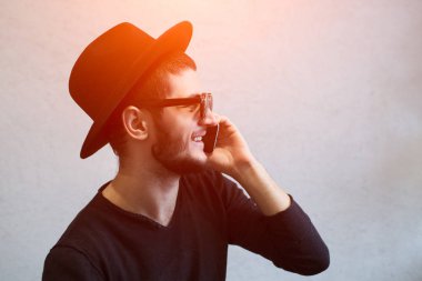 Genç adam güneş gözlüğü ve siyah şapka giymiş, beyaz arka plan üzerinde akıllı telefon kullanan gülümseyen portresi