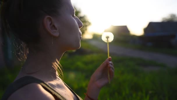 Μεγέθυνση της εφηβικής ηλικίας κορίτσι φυσάει πικραλίδα στο ηλιοβασίλεμα. — Αρχείο Βίντεο